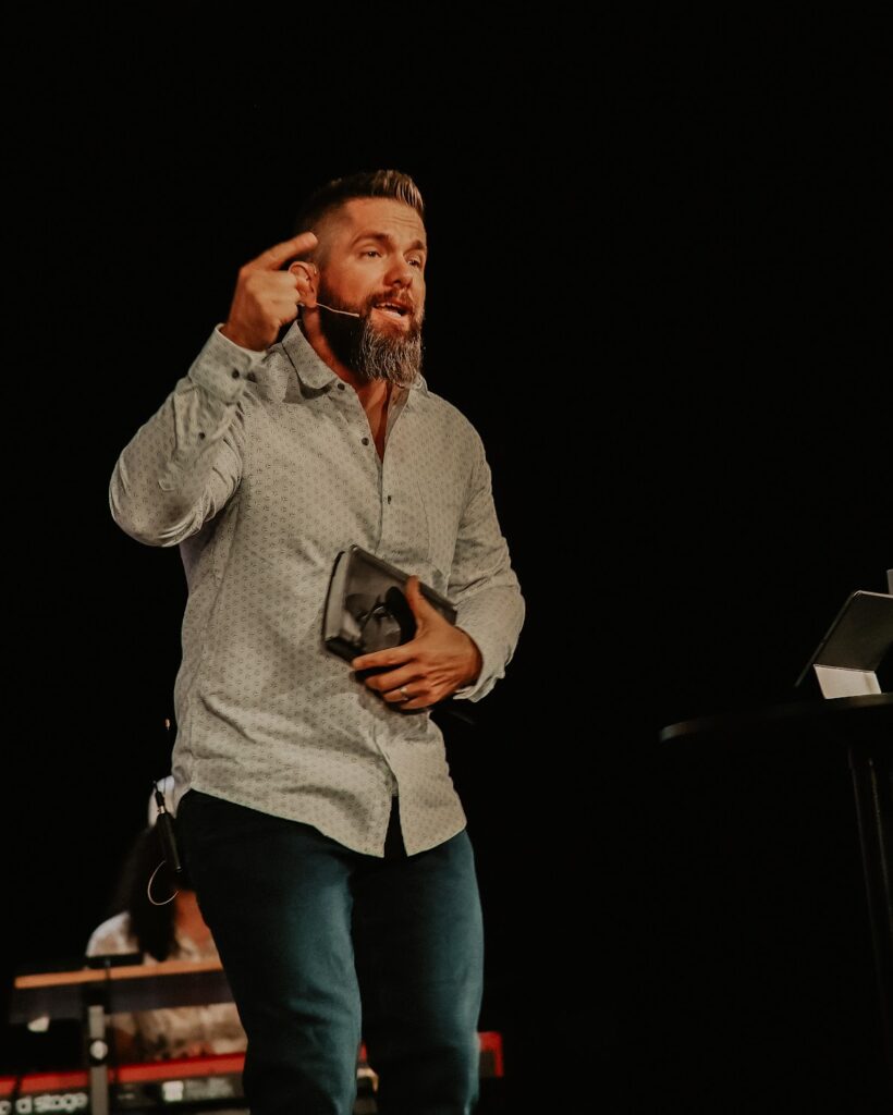 Rick Scadden stands with his right hand raised, pointed to the sky, and a Bible in his left hand