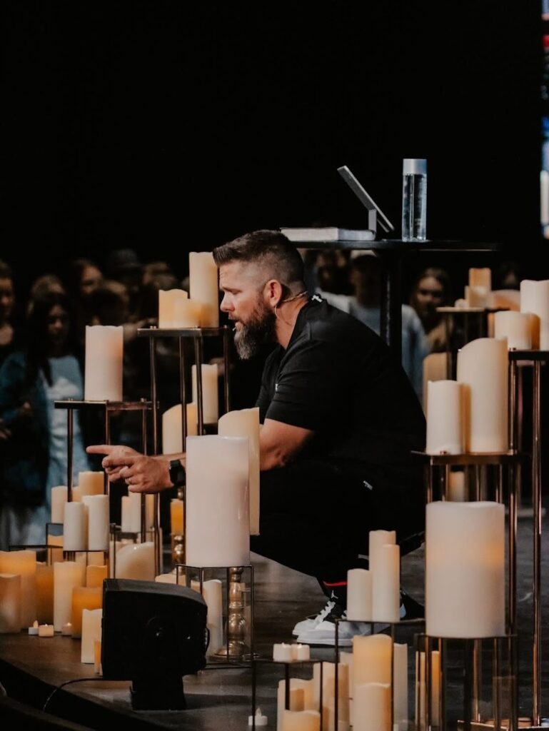 Rick Scadden kneels in prayer in front of a crowd in a dark room surrounded by white candles