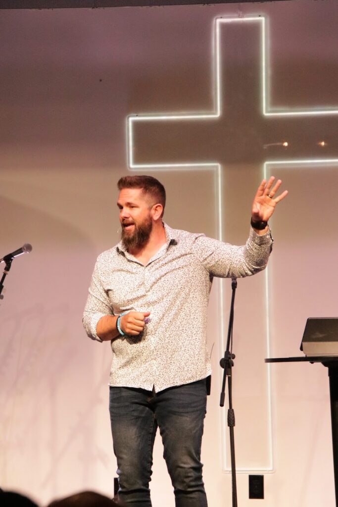 Rick Scaddon, a man with a beard, white button down shirt and jeans stands on a stage with a white neon cross sign in the background